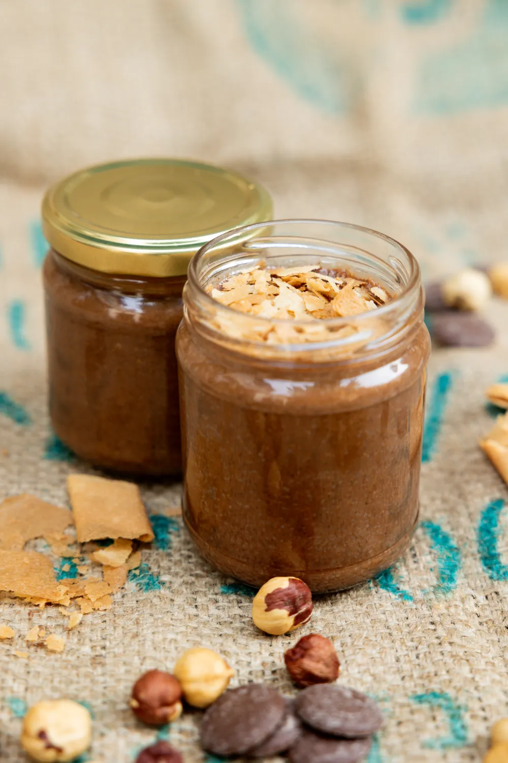 Deux pots de pâte à tartiner au chocolat, l'un garni d'éclats de pralin, entourés de noisettes et de palets de chocolat